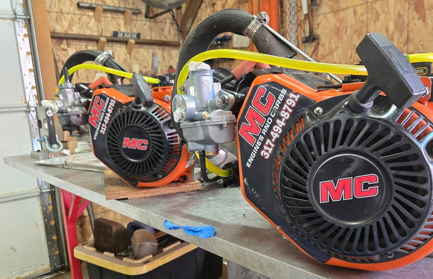 Two kart lo206 engines sitting on aluminum bench top, in a garage with some wrenches hanging int he background. The lo206 engines are made by briggs and stratton and are orange on the outside.