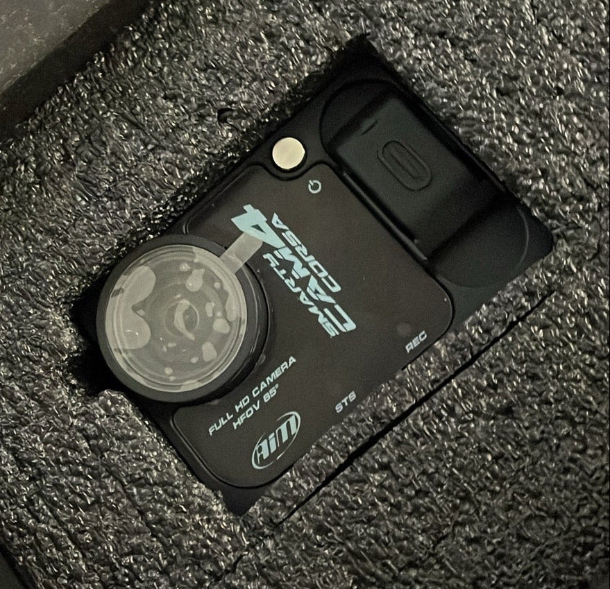A smarty cam 4 corsa inside of foam packaging , the camera is black and has a plastic lens protection cover, the foam surrounding the camera is black.