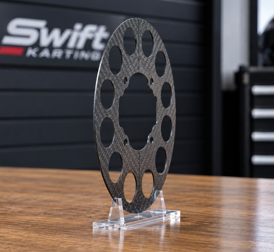 A carbon fiber smoker performance guard guard with a visible carbon weave pattern, displayed on a clear acrylic stand on a wooden table with a blurred Swift Karting background.